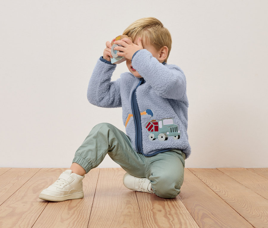 Ein blonder Junge kniet auf einem Holzboden und hält eine Spielzeugkamera vor sein Gesicht. Er trägt eine blaue Fleecejacke mit einem Lastwagen-Applikation und grüne Hosen.