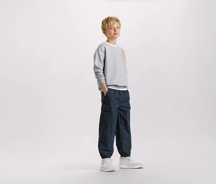 Ein blonder Junge steht mit den Händen in den Taschen in einem grauen Sweatshirt und marineblauen Cargohosen.