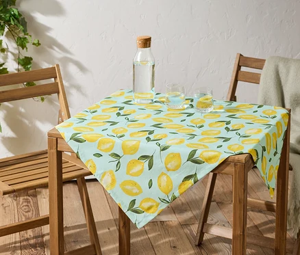 Ein Tisch mit einer hellblauen Tischdecke mit Zitronenmuster steht zwischen zwei Holzstühlen. Auf dem Tisch stehen eine Wasserflasche und zwei Gläser Wasser.