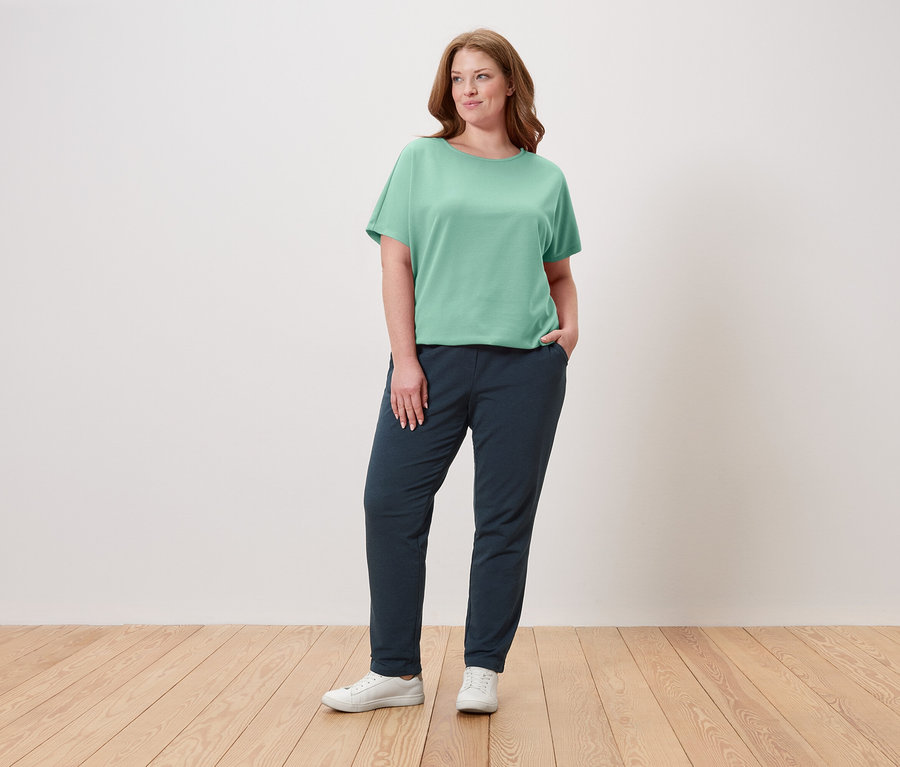 Eine Frau steht mit der Hand in der Hosentasche und trägt ein grünes T-Shirt, eine marineblaue Hose und weiße Turnschuhe.