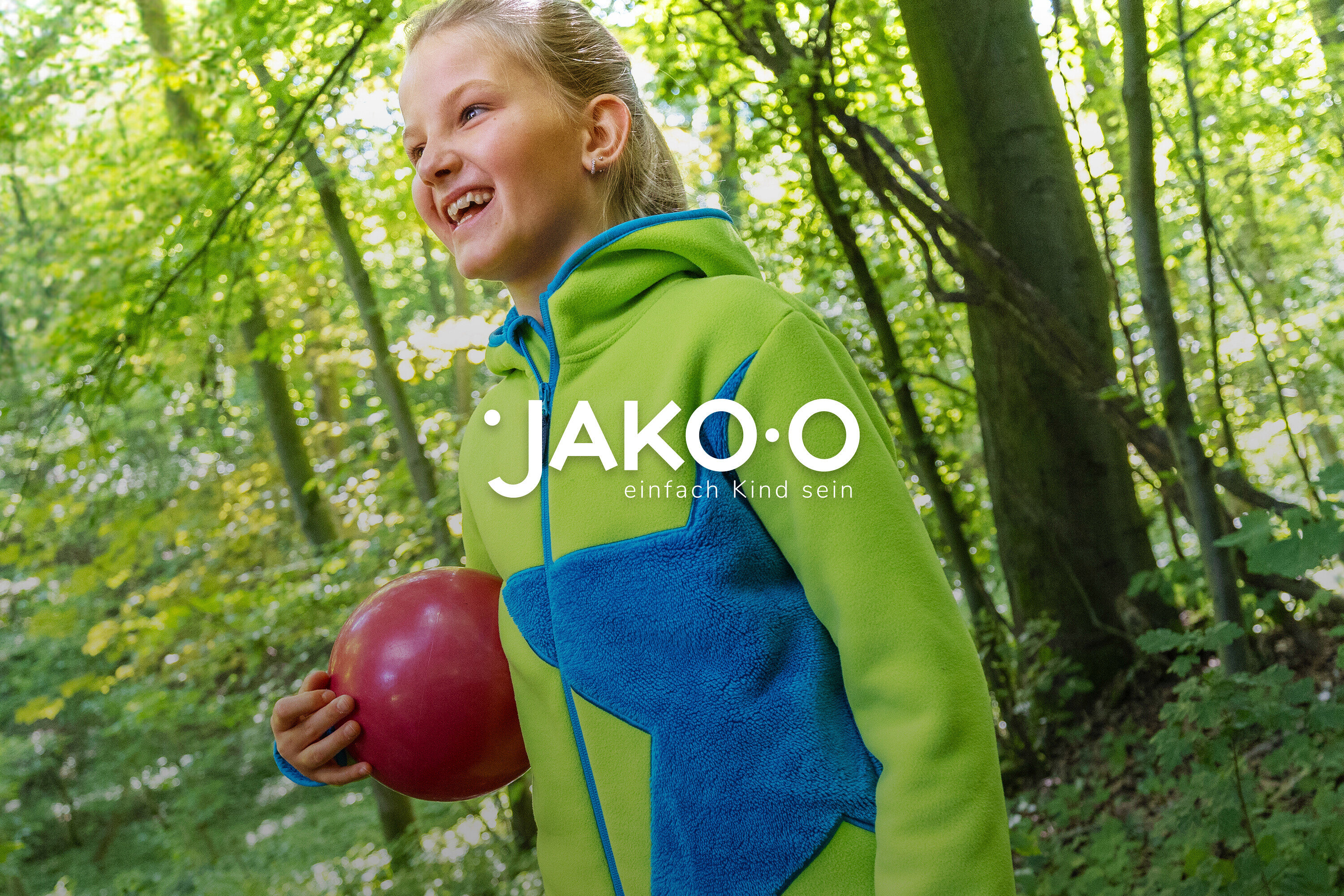 Lachendes Mädchen mit rotem Ball in grün-blauer JAKO-O Jacke im Wald.