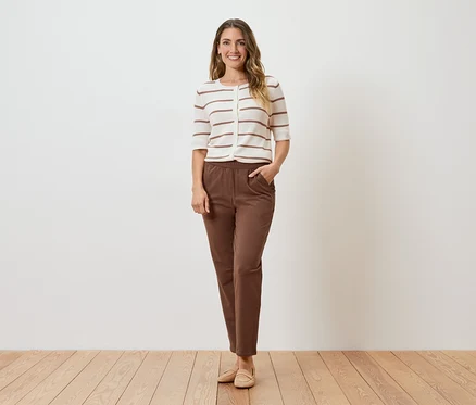 Eine Frau steht vor einer weißen Wand und trägt eine gestreifte Strickjacke, braune Hosen und beigefarbene Schuhe.