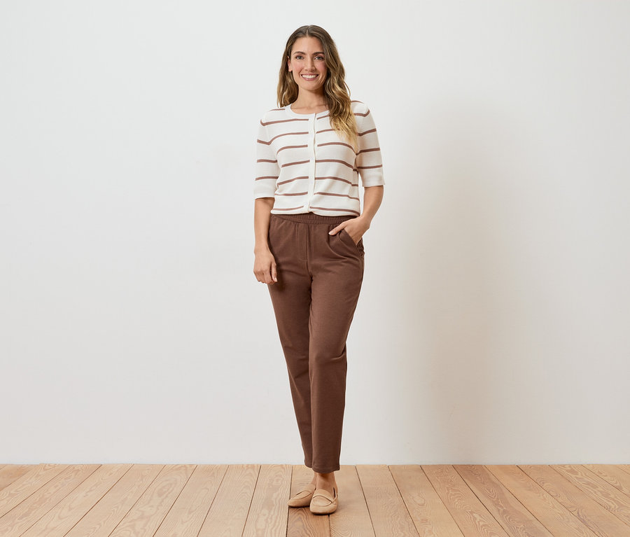 Eine Frau steht vor einer weißen Wand und trägt eine gestreifte Strickjacke, braune Hosen und beigefarbene Schuhe.