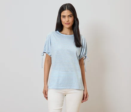 Frau mit blau-weiß gestreiftem T-Shirt mit kurzen Ärmeln und beigen Hosen vor einem hellen Hintergrund.
