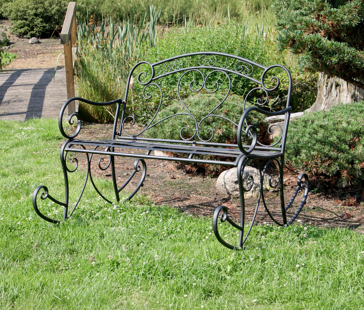 Eine schwarze Metall-Gartenbank steht auf einer Rasenfläche vor Büschen und einem Baum.