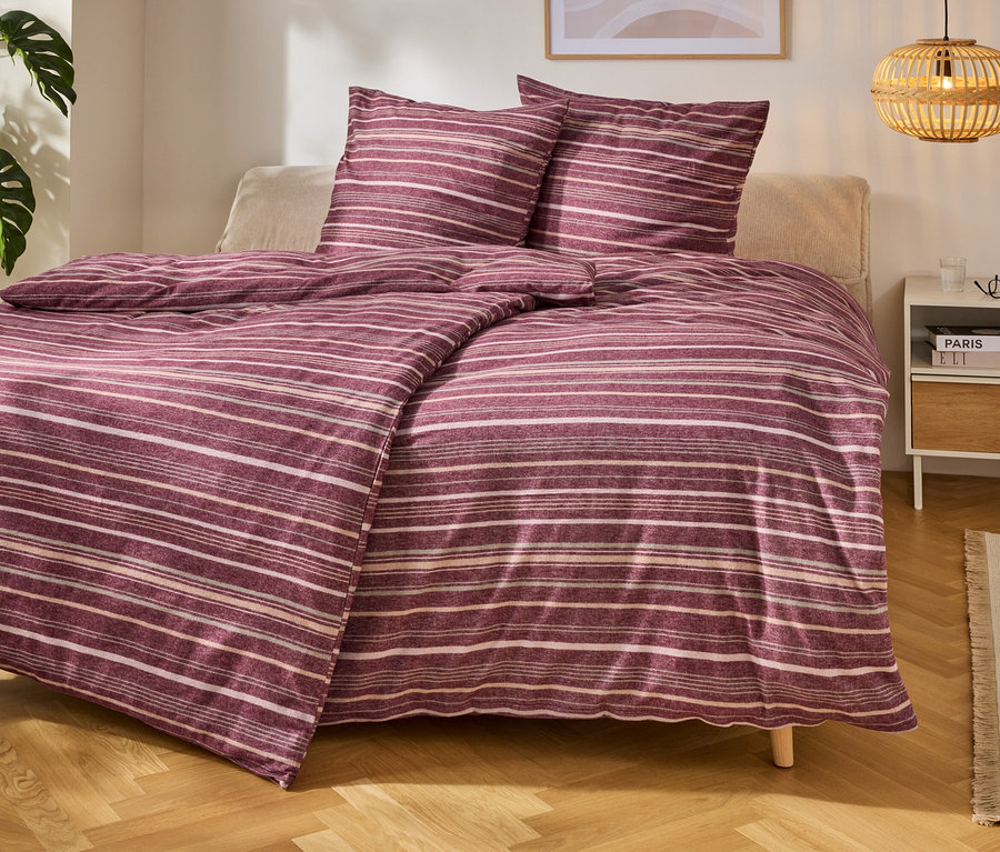 Ein Bett mit gestreifter Bettwäsche in Lila- und Weißtönen, daneben ein Nachttisch mit Büchern und einer Lampe. Links steht eine Pflanze.