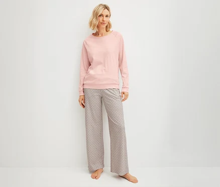 Blonde Frau in pinkem Langarmshirt und gemusterter CALIDA »Favorites Arts« Hose steht barfuß vor weißer Wand.
