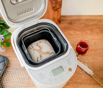 Ein Brotbackautomat mit geöffnetem Deckel zeigt ein frisch gebackenes Brot. Daneben stehen ein Glas Marmelade und ein Messer auf einer Holzoberfläche.