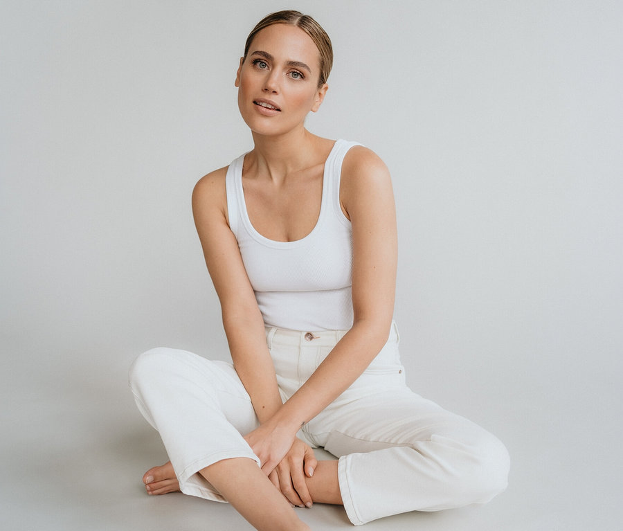 Eine Frau sitzt barfuß im Schneidersitz auf dem Boden und trägt ein weißes Tanktop und weiße Angels Denim »Evy Cool« O-Shape Jeans.