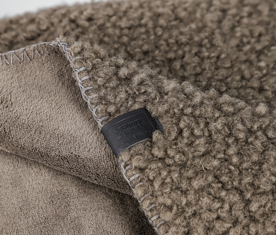 Nahaufnahme einer zweifarbigen Decke. Eine Seite ist flauschig und zottelig in Dunkelbeige, die andere Seite ist glatt und hellbeige. Der Rand ist mit hellgrauen Zickzacknähten versehen. Ein dunkles Etikett mit der Aufschrift 'SCHÖNER WOHNEN KOLLEKTION' ist auf der flauschigen Seite angenäht.