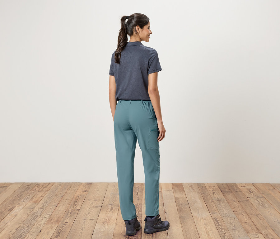 Rückansicht einer Frau mit Pferdeschwanz, die ein marineblaues Poloshirt, grüne Hosen und Wanderschuhe vor einer weißen Wand und einem Holzboden trägt.