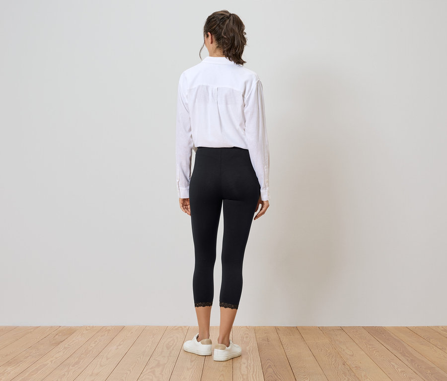 Rückansicht einer Frau mit Pferdeschwanz, die eine weiße Bluse, schwarze Leggings mit Spitzenbesatz und weiße Turnschuhe auf einem Holzboden trägt.