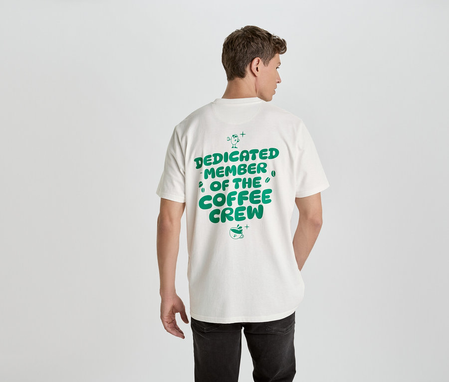 Ein Mann von hinten trägt ein weißes T-Shirt mit grünem Aufdruck 'Dedicated Member of the Coffee Crew'.