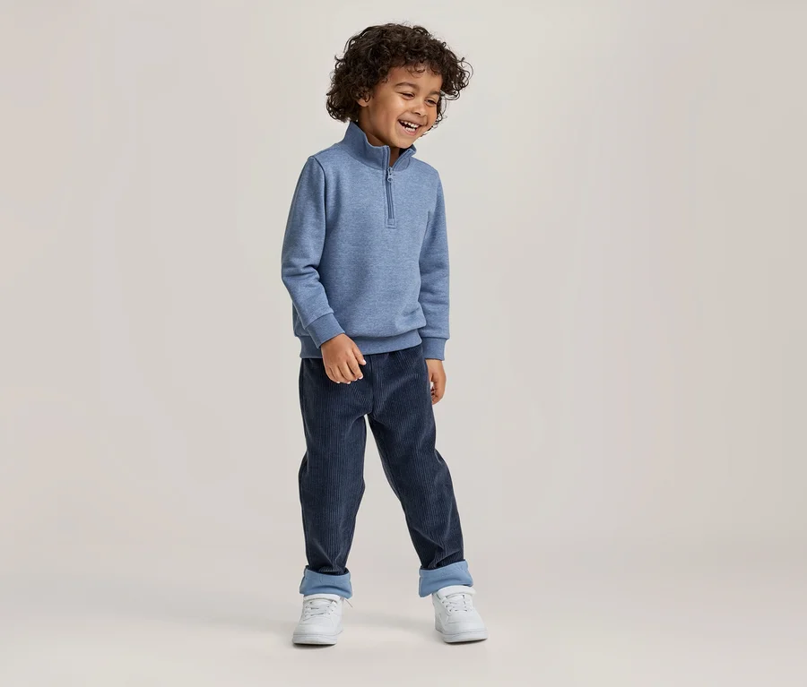 Ein lächelnder Junge mit lockigem Haar trägt ein blaues Sweatshirt mit Reißverschluss, dunkelblaue Cordhosen mit umgeschlagenen Bündchen und weiße Turnschuhe.