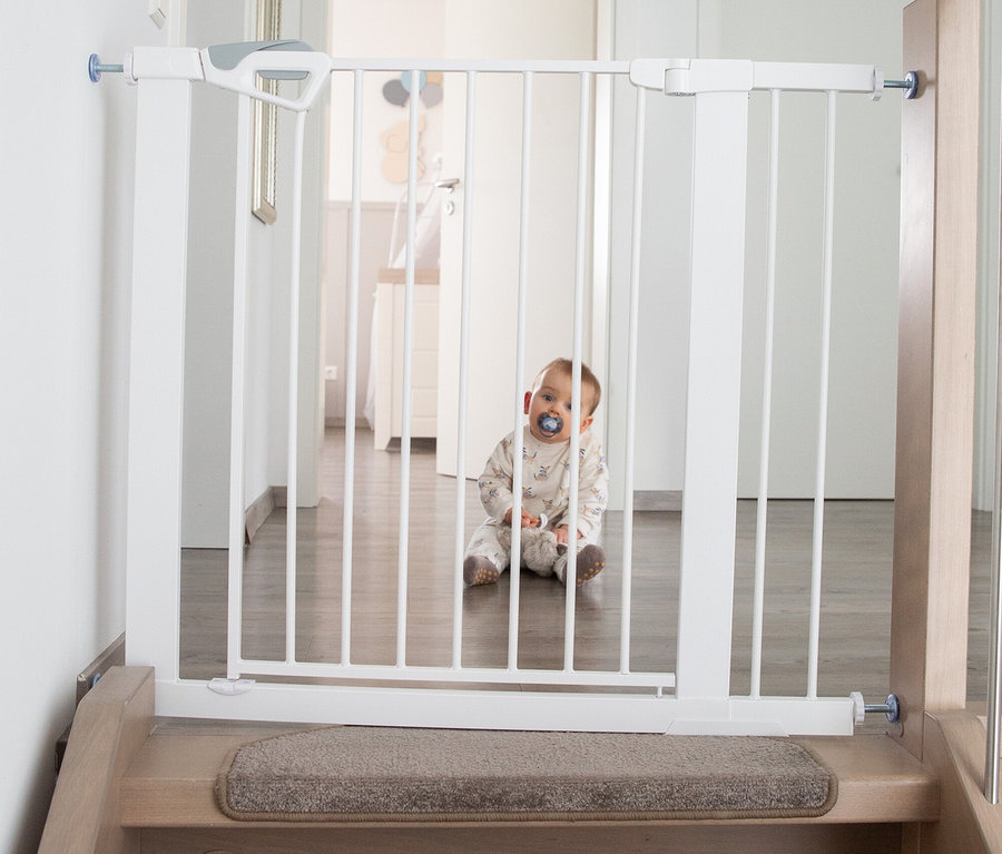 Ein Baby mit Schnuller sitzt hinter einem weißen roba easySafe+ Tür- und Treppenschutzgitter, das oben an einer Holztreppe einen Flur absperrt.