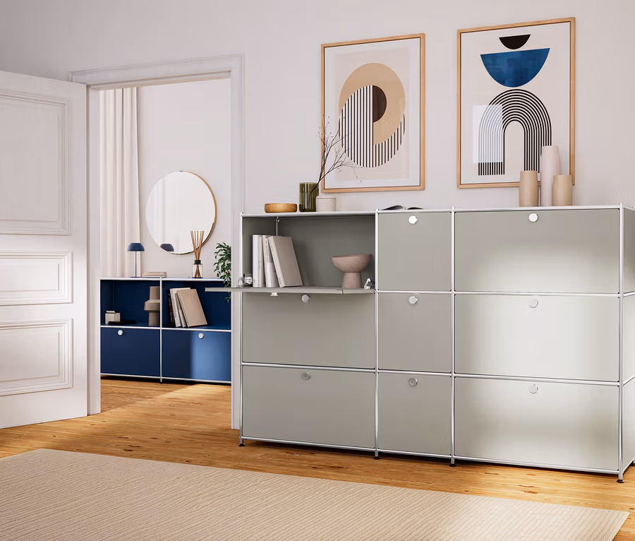 Helles Wohnzimmer mit beigem Teppich und einem großen, hellgrauen modularen Aufbewahrungssystem. Darüber hängen zwei gerahmte, abstrakte Bilder. Durch eine offene weiße Tür ist ein weiterer Raum mit einem dunkelblauen Aufbewahrungssystem und einem runden Spiegel sichtbar.
