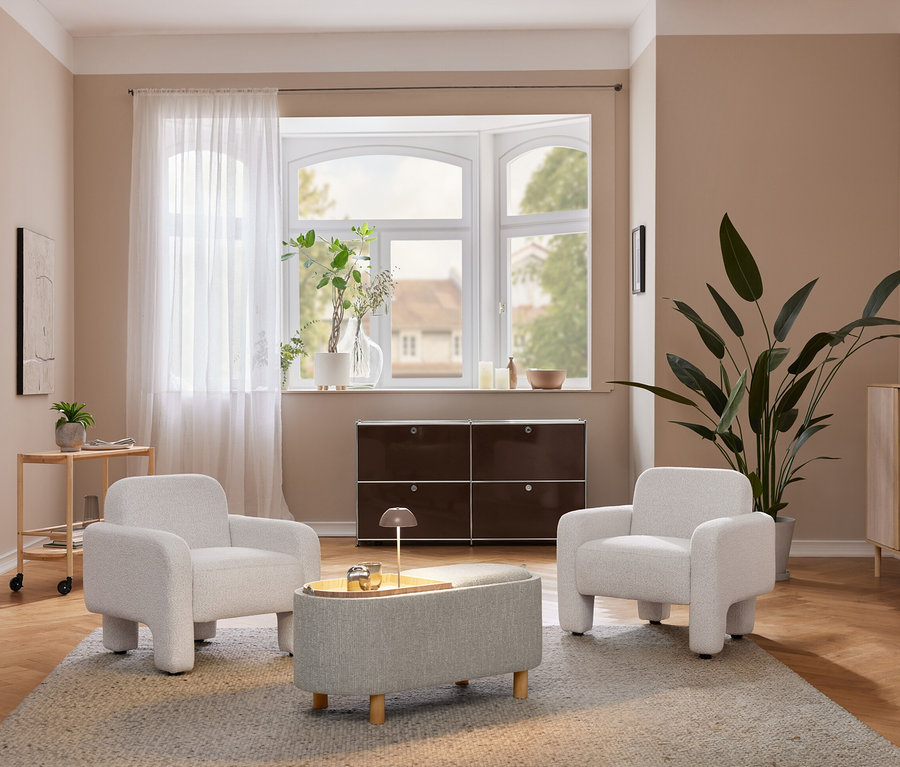 Gemütliches Wohnzimmer mit zwei hellgrauen Sesseln und einem passenden Ottoman auf einem Teppich. Eine beige LED-Akku-Tischleuchte steht auf einem hellen Holzschrank neben einer großen Pflanze. Unter einem Erkerfenster befindet sich ein dunkelbraunes Aufbewahrungsmöbel.
