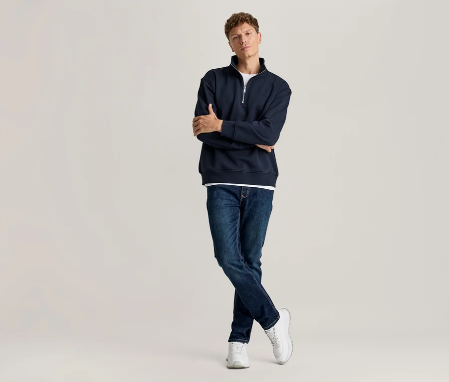 Mann steht mit verschränkten Armen, trägt ein dunkelblaues Half-Zip-Sweatshirt über einem weißen T-Shirt, blaue Jeans und weiße Sneaker.