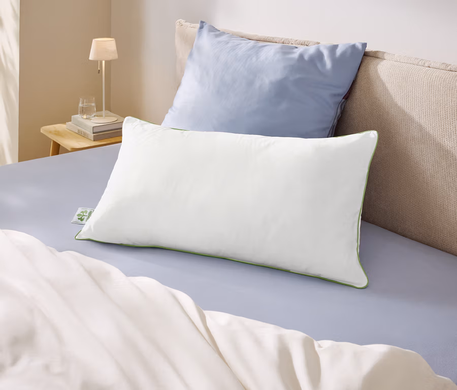 Ein Bett mit hellblauem Laken, einer weißen Decke und zwei Kissen. Ein weißes Kissen mit grünem Rand liegt vor einem hellblauen Kissen. Links daneben steht ein Nachttisch mit Lampe und Wasserglas.