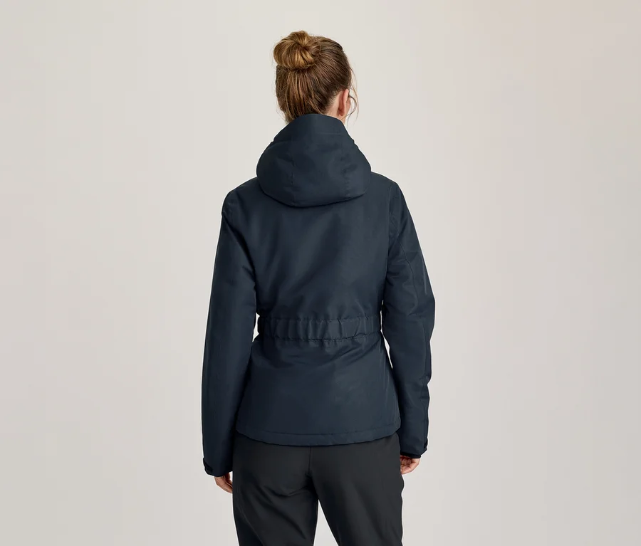 Frau von hinten, trägt eine dunkelblaue Kapuzenjacke mit verstellbarer Taille und schwarze Hose. Ihr Haar ist zu einem Dutt gebunden.