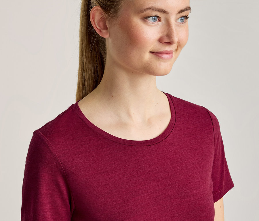 Junge Frau mit blondem Pferdeschwanz trägt ein weinrotes T-Shirt mit Rundhalsausschnitt und blickt lächelnd nach rechts.