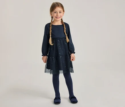 Lächelndes Mädchen in dunkelblauem, langärmeligem Kleid mit glitzerndem Tüllrock, dunkelblauer Strumpfhose und Samtschuhen. Sie trägt zwei Zöpfe.
