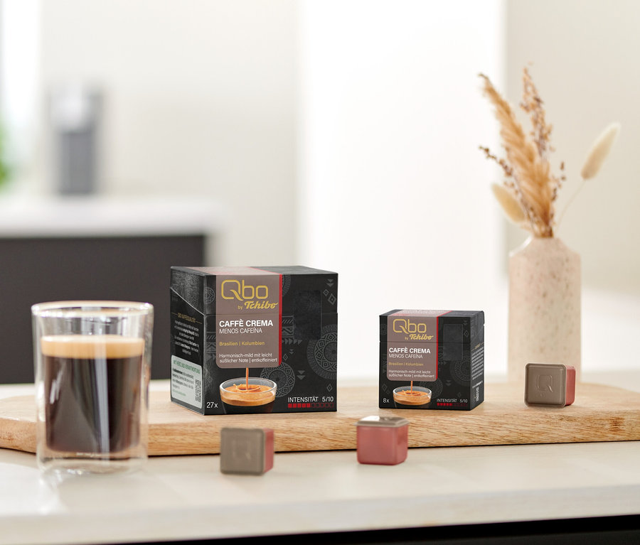 Auf einem Tisch stehen Qbo Caffè MENOS CAFEÍNA Kapselpackungen und eine Tasse Kaffee auf einem Holzbrett.