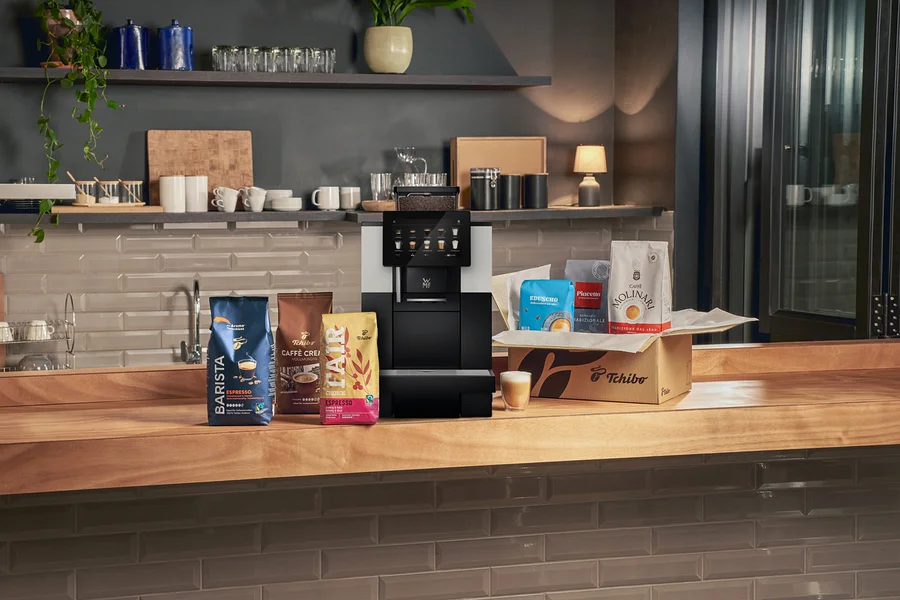 Eine schwarz-silberne Kaffeemaschine steht auf einer Holztheke, umgeben von mehreren Kaffeebohnenbeuteln. Links liegen blaue, braune und gelbe Kaffeebeutel. Rechts befindet sich eine Tchibo-Box mit drei weiteren Kaffeebeuteln und davor ein kleines Glas mit geschichtetem Kaffee.