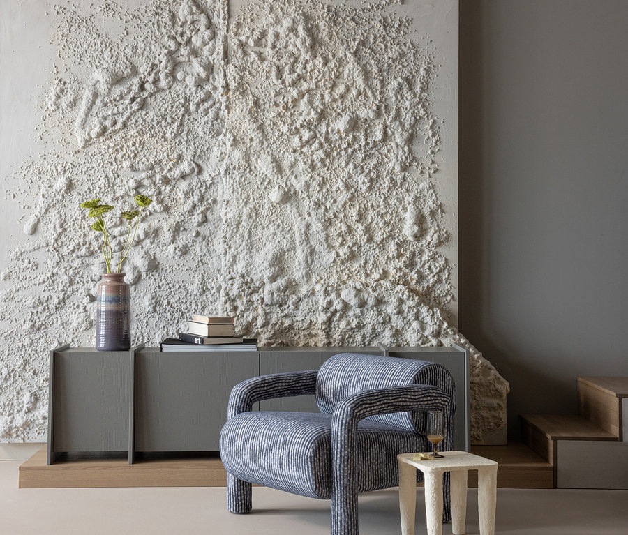 Ein modernes Zimmer mit einem blau-weiß gestreiften Sessel neben einem strukturierten weißen Beistelltisch mit einem Getränk. Dahinter steht ein graues Sideboard mit einer Vase mit Pflanzen und Büchern vor einer stark strukturierten weißen Wand. Rechts sind Holzstufen sichtbar.