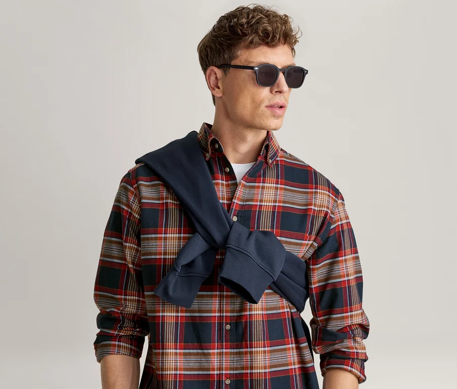 Mann in rot-blau-weißem Karomusterhemd, schwarzer Sonnenbrille und dunkelblauem Sweatshirt um die Schultern gebunden.