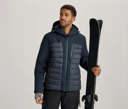 Mann in dunkelblauer und grauer Skijacke und schwarzer Skihose, der ein Paar schwarze Skier hält.