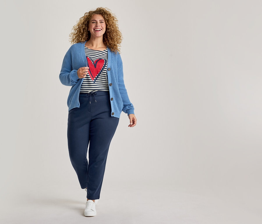 Eine lächelnde Frau mit lockigem Haar trägt einen blauen Strickcardigan, ein gestreiftes T-Shirt mit rotem Herz, dunkelblaue Jogginghose und weiße Sneaker, während sie nach vorne geht.