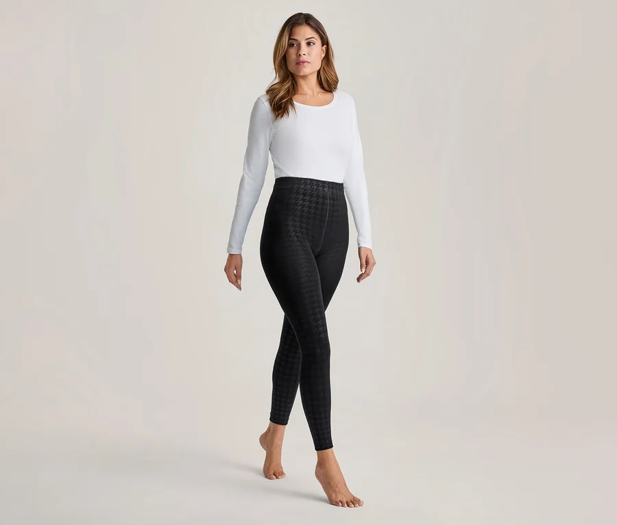 Frau in weißem Langarmshirt und schwarzen Leggings mit Hahnentrittmuster, barfuß stehend.