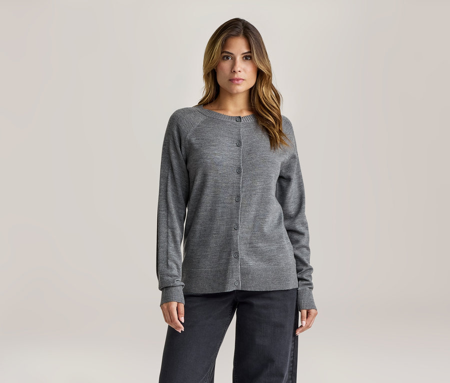 Frau mit mittellangem braunem Haar trägt einen grauen Strickcardigan mit Knöpfen und eine dunkle weite Hose, blickt nach vorne.