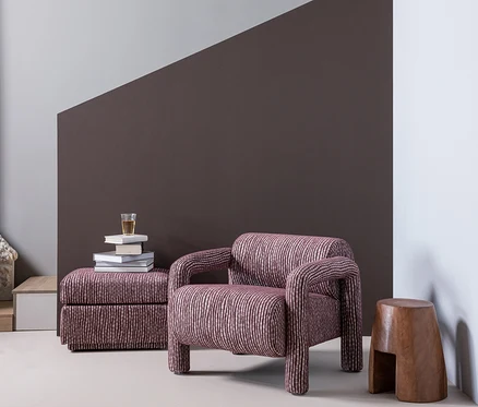 Ein gestreifter Sessel und ein passender Hocker in einem Raum mit einer diagonal geteilten Wand in Grau und Dunkelbraun. Auf dem Hocker liegen Bücher und ein Glas, daneben steht ein Holzschemel.