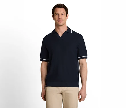 Ein Mann steht frontal da und trägt ein dunkelblaues, kurzärmeliges Poloshirt mit weißen Streifen am Kragen und an den Ärmeln sowie eine hellbraune Hose.