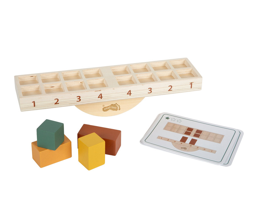 Eine hölzerne Waage mit nummerierten Schlitzen, bunten Holzklötzen und Anleitungskarten. Die helle Holzwaage hat auf jeder Seite die Zahlen 1 bis 4 mit quadratischen Schlitzen darüber. Daneben liegen vier Klötze in Grün, Orange, Gelb und Braun sowie zwei Karten mit Anwendungsdiagrammen.
