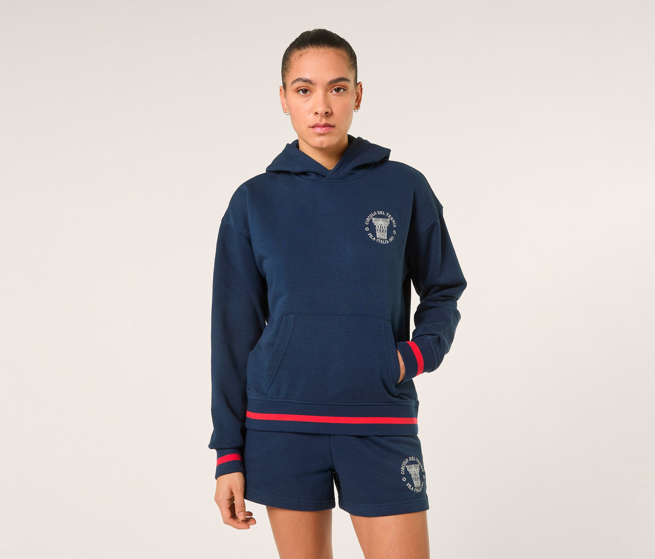 Eine Frau trägt einen dunkelblauen FILA Damen-Hoodie »MARLIA« mit weißem Logo und roten Streifen an den Bündchen, dazu passende dunkelblaue Shorts mit Logo und rotem Saum.