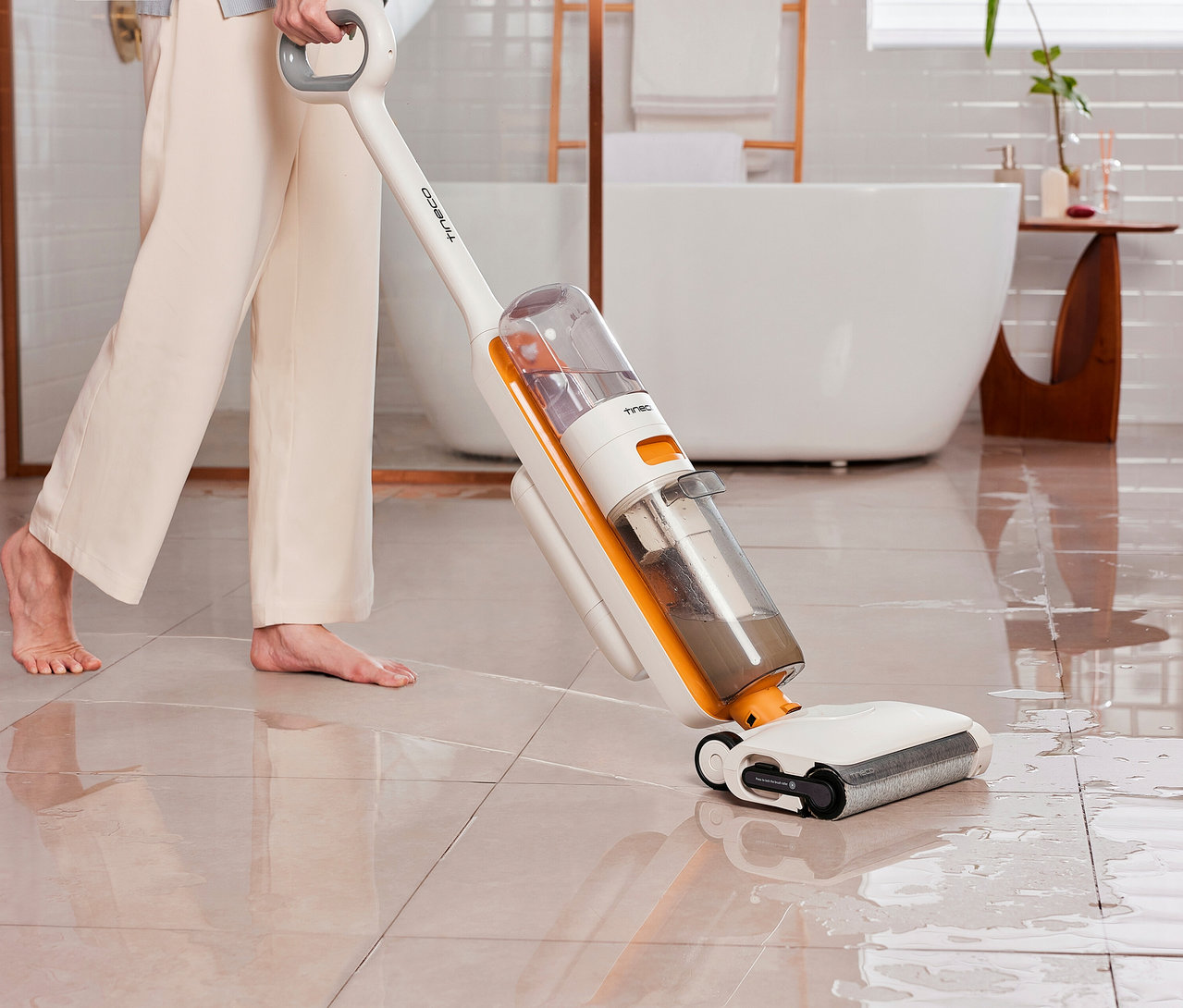 Eine barfüßige Person in heller Hose reinigt mit dem weißen und orangefarbenen Tineco 2-in-1-Nass-Trockensauger »Y2 Plus« einen nassen Fliesenboden im Badezimmer. Der Sauger hat sichtbare Wassertanks, einer davon mit Schmutzwasser.