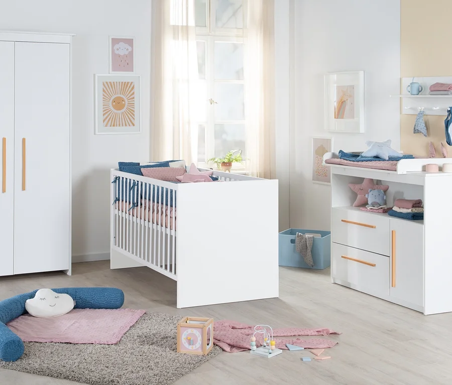 Weißer Kleiderschrank mit Holzgriffen, weißes Gitterbett mit rosa und blauen Kissen, Wickelkommode mit Wickelauflage und Regalen. Auf dem Boden liegt ein Teppich mit einer blauen Nackenrolle und einem Wolkenkissen. An der Wand hängen Bilder und über der Wickelkommode ein Regal mit Babyartikeln.