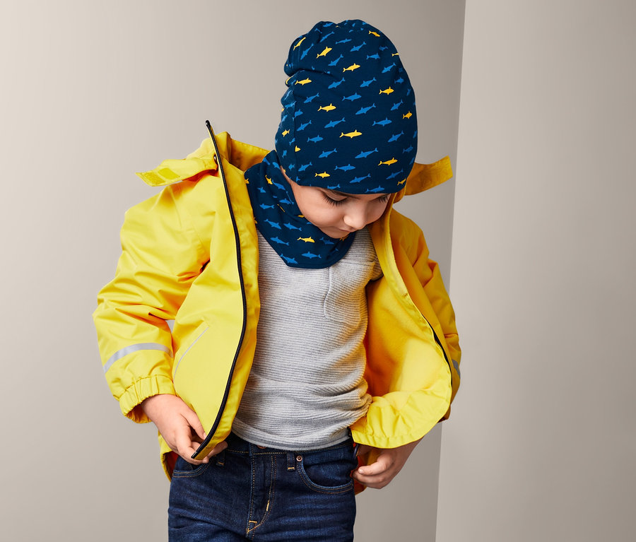 Junge mit gelber Jacke, der den Reißverschluss seiner Jacke richtet und eine Mütze und ein Halstuch trägt.
