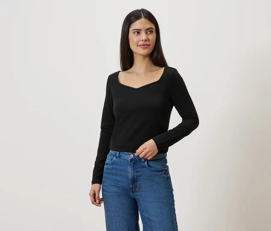 Frau mit langen dunklen Haaren trägt ein schwarzes Langarmshirt und blaue Jeans vor einem weißen Hintergrund.