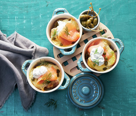 Drei antiktürkise STAUB Keramik-Cocottes mit Kartoffelgericht, Lachs, Sauerrahm und Dill auf Holztrivet. Daneben ein Deckel und Kapernbeeren.