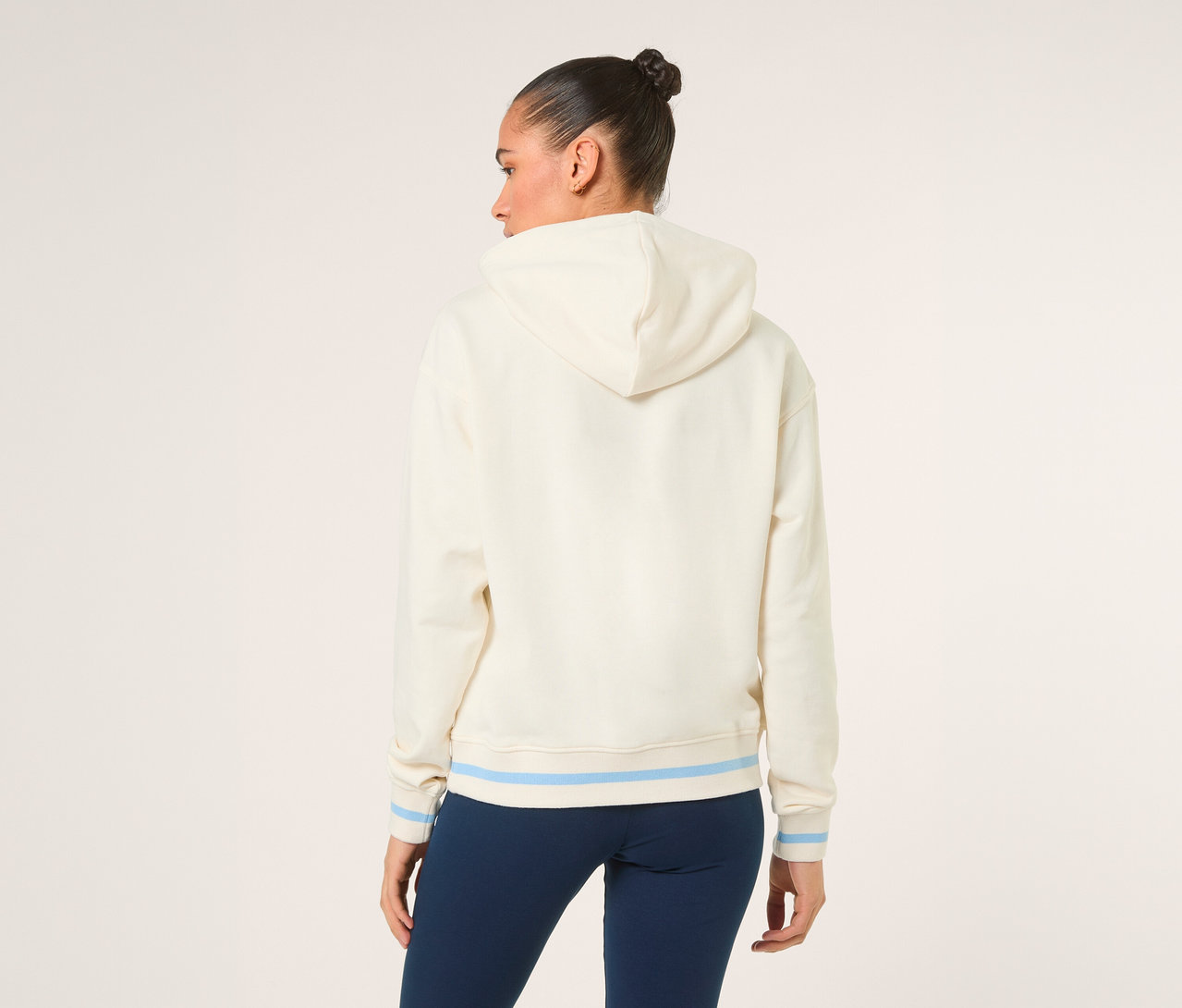 Frau von hinten, trägt einen cremefarbenen FILA Damen-Hoodie »MARLIA« mit hellblauen Streifen an Saum und Ärmelbündchen sowie dunkelblaue Leggings.