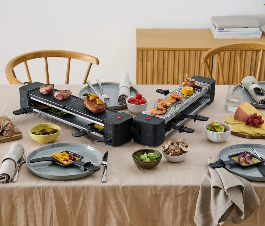 Zwei klappbare Raclette-Grills mit Speisen auf einem Tisch.