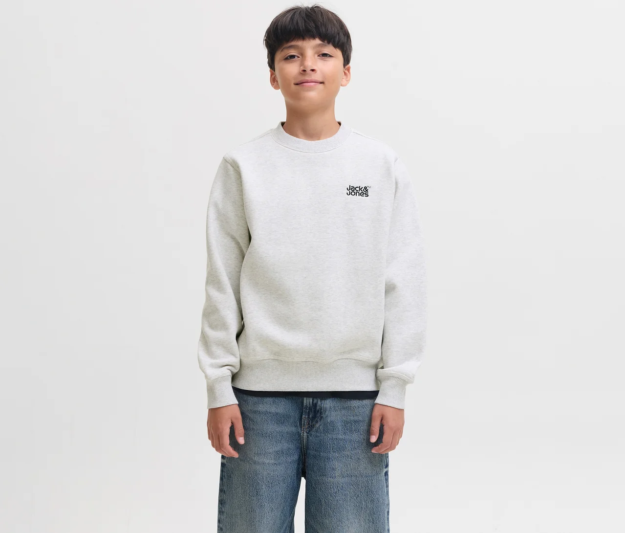 Ein junger Junge mit dunklen Haaren lächelt leicht und trägt einen weißen Jack&Jones Sweater Colton mit dem Markenlogo auf der Brust und blaue Jeans.
