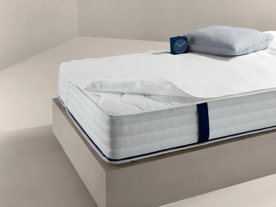 Bett mit weißem Matratzenschoner, ca. 140 x 200 cm, Kissen und Uhr.