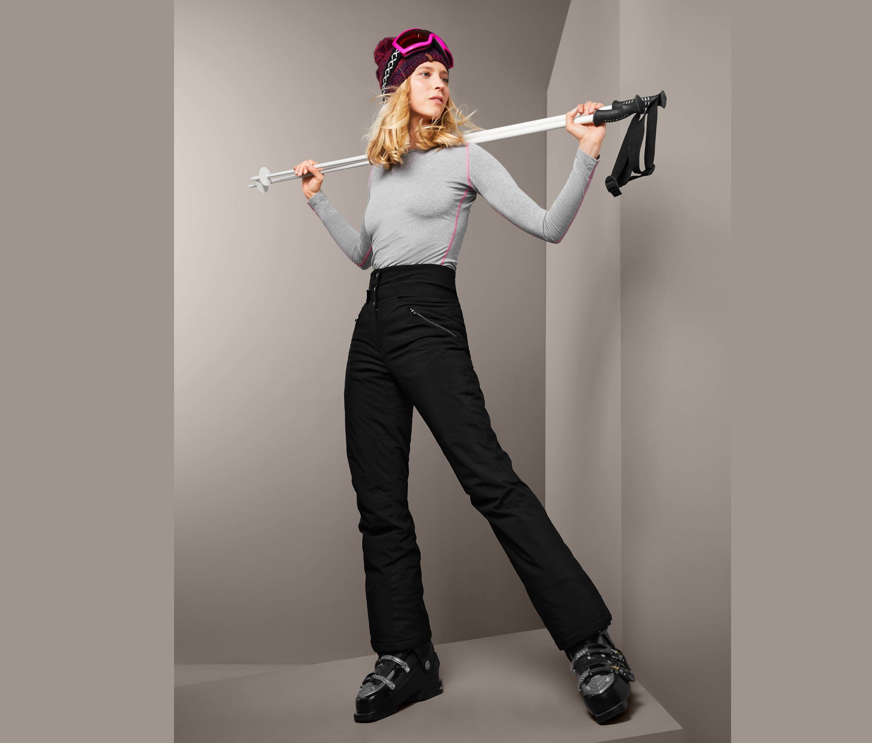 Blonde Frau in grauem Shirt und schwarzer High-Waist-Softshell-Skihose hält Skistöcke.