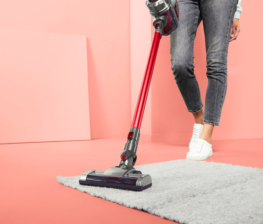 Eine Frau saugt mit einem Akkusauger einen grauen Teppich in einem Raum mit korallenfarbenen Wänden.
