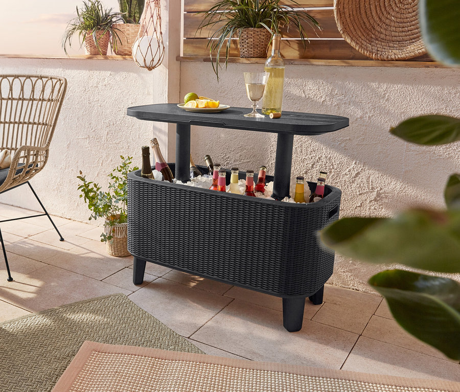Terrasse mit rundem Tisch, Boho-Stühlen, Outdoor-Teppich und einer Coolbar voller Getränke. Eine geflochtene LED-Hängeleuchte hängt über dem Tisch und eine LED-Laterne steht in der Nähe.